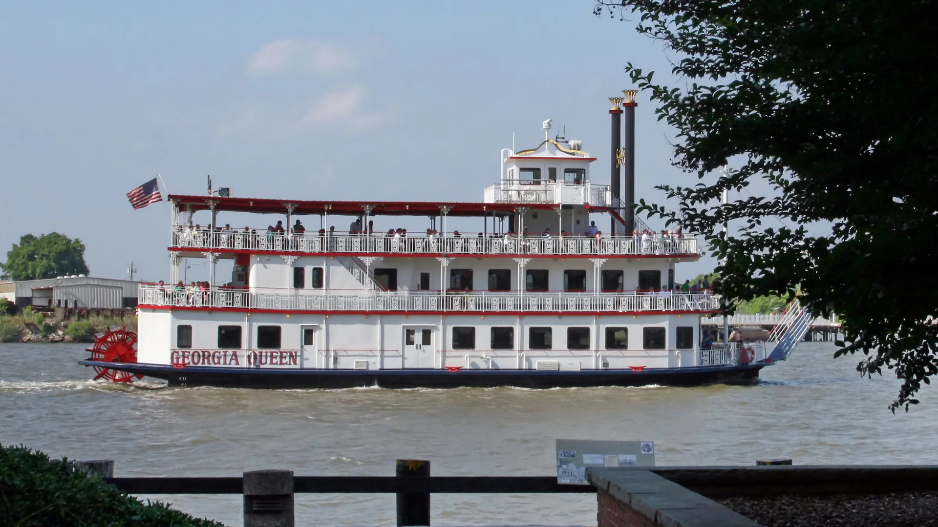 A white paddlewheel riverboat with red trim and two black stacks moving along the Savannah River, with the red paddlewheel at its stern and a dark tree framing the right edge.