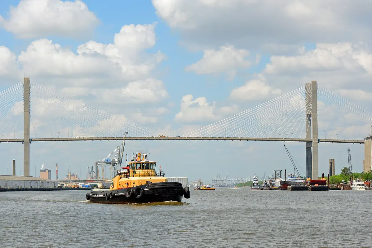 A yellow and black tug on the Savannah River in front of the twin-towered Talmadge Memorial Bridge under a cloudy blue sky.