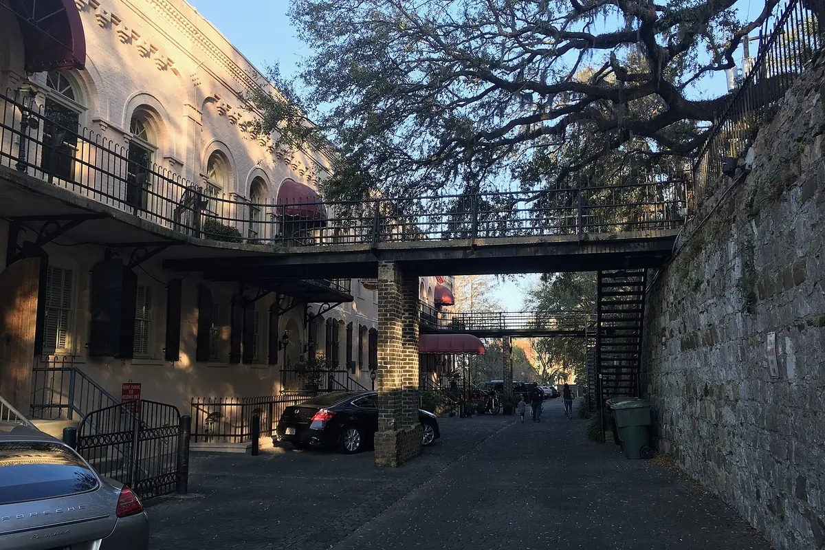 A footbridge and a moss-draped live oak above a cobbled ramp at Factors Walk, beside a tall stone retaining wall.