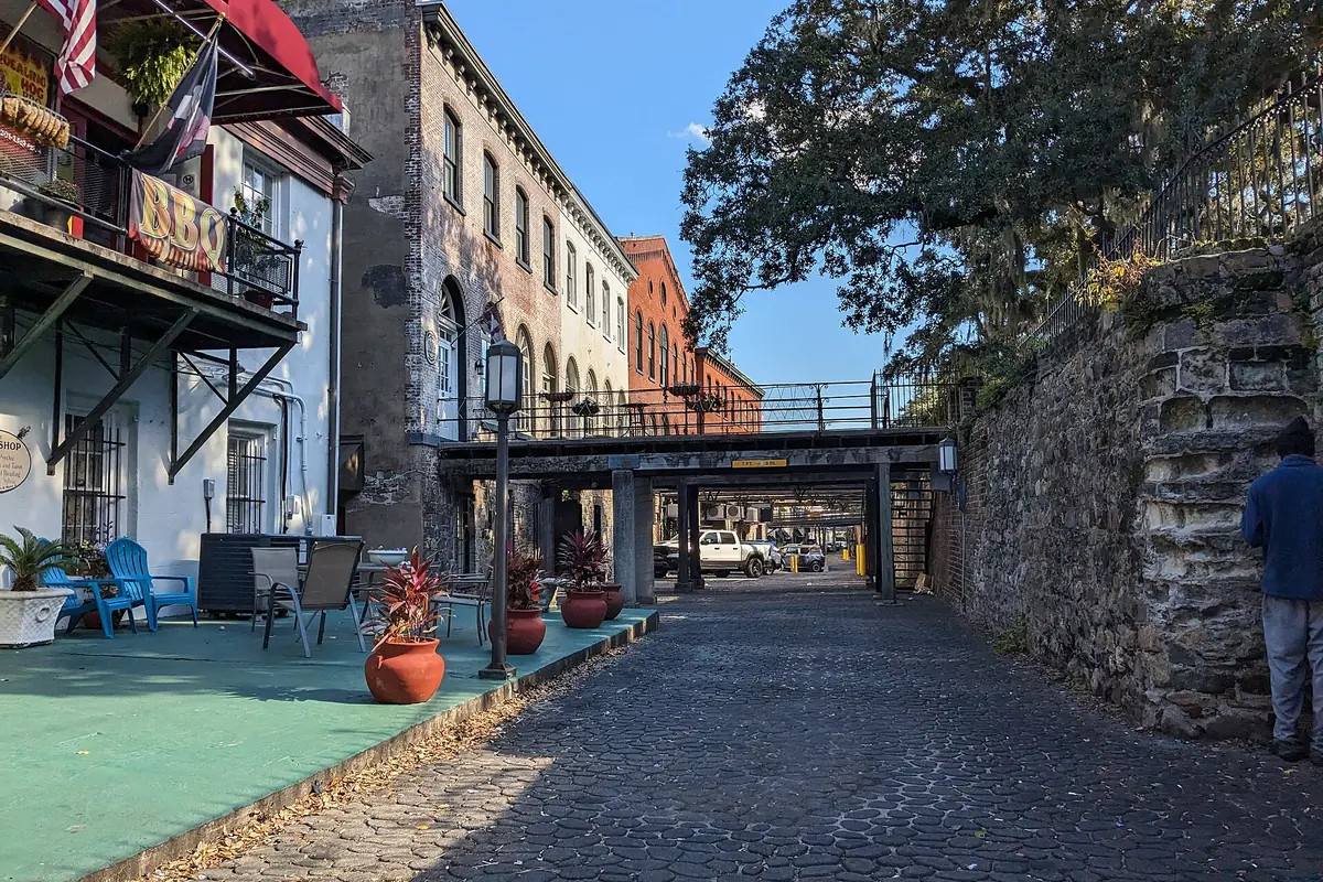 A cobbled ramp passing under a Factors Walk footbridge, between old brick warehouses and a stone wall hung with moss.
