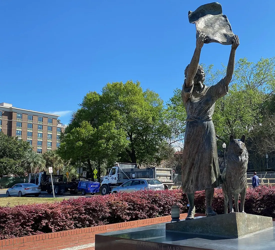 A bronze statue of a woman waving a cloth with a dog at her side, on a brick plinth with a red hedge